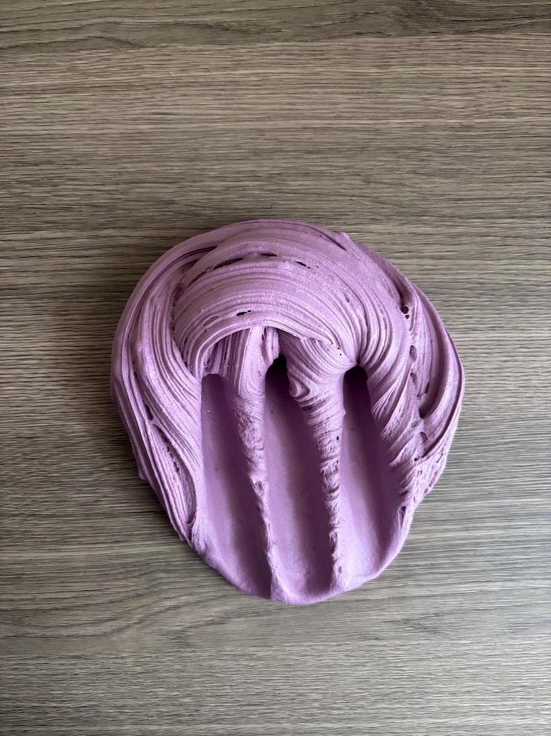 May include: A mound of light purple slime with a thick, textured consistency. The surface features deep, swirling ridges and elongated indentations that create a layered, malleable appearance across the entire circular-shaped mass of material.