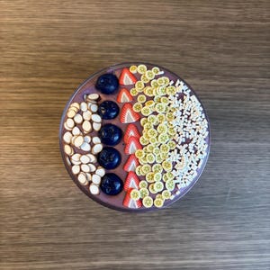 May include: A round bowl filled with a purple smoothie base topped with rows of dark blue blueberries, white almond slices, red strawberry slices, small yellow kiwi slices, and white rectangular sprinkles arranged in a decorative pattern.