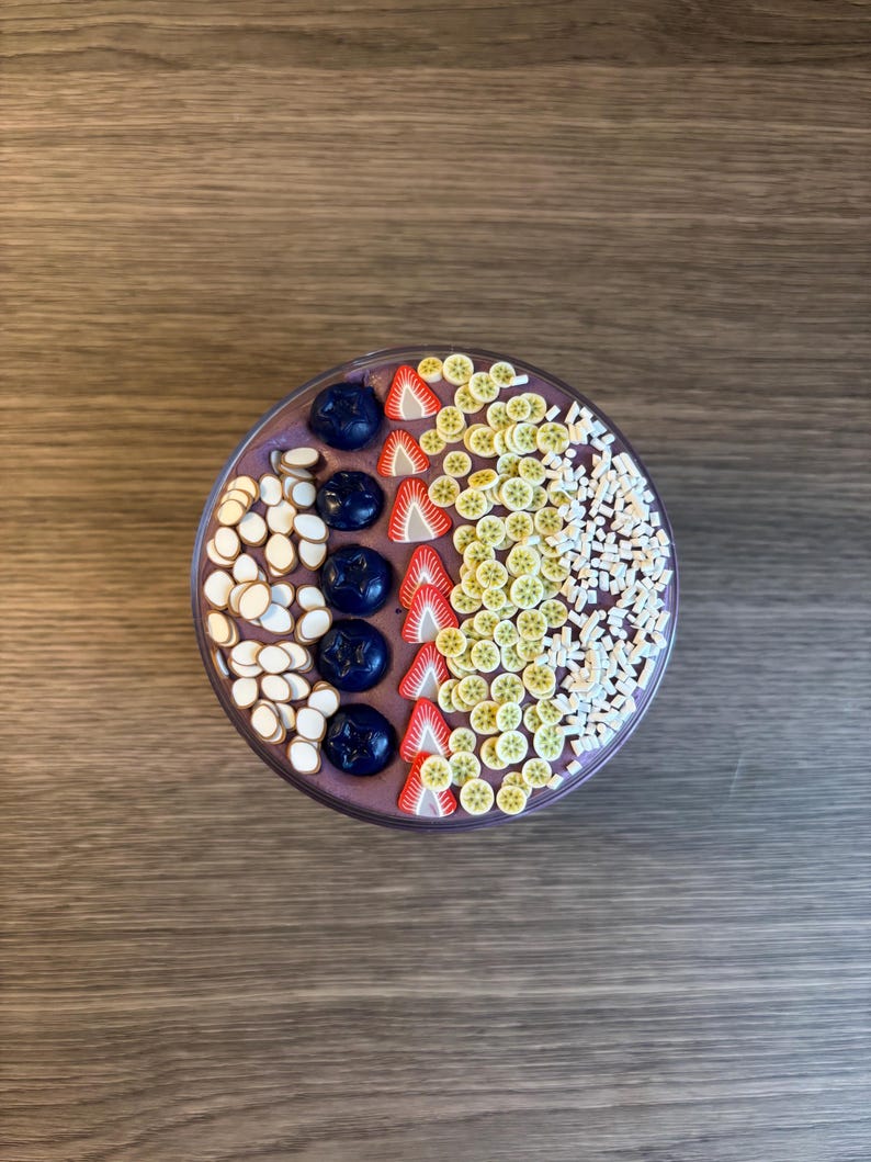 May include: A round bowl filled with a purple smoothie base topped with rows of dark blue blueberries, white almond slices, red strawberry slices, small yellow kiwi slices, and white rectangular sprinkles arranged in a decorative pattern.