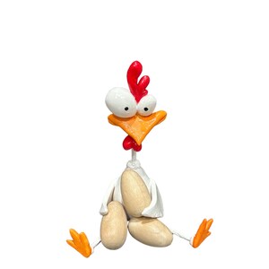 May include: A white ceramic chicken figurine with red and orange accents, holding three beige eggs. The chicken has large, white eyes and a comical expression.