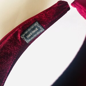 May include: A close-up of a burgundy velvet headband with a black label that reads "HAND MADE".