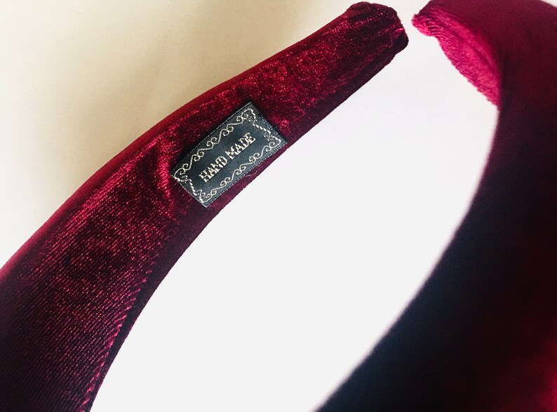 May include: A close-up of a burgundy velvet headband with a black label that reads "HAND MADE".