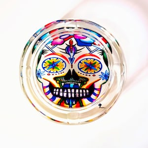 Puede incluir: Un cenicero de vidrio transparente con un diseño de calavera de azúcar colorido. La calavera tiene detalles brillantes en amarillo, azul, rojo y verde.