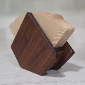 May include: A dark brown wooden coffee filter holder with a hexagonal shape, containing a stack of light tan, unbleached paper coffee filters. The holder features a solid wood construction with a smooth finish and angled sides to support the filters.