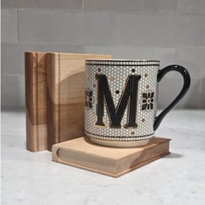 May include: A ceramic mug featuring a black letter M on a white honeycomb pattern with gold accents and a black handle, positioned next to and resting upon light-colored wooden blocks that resemble the spines and covers of small books.