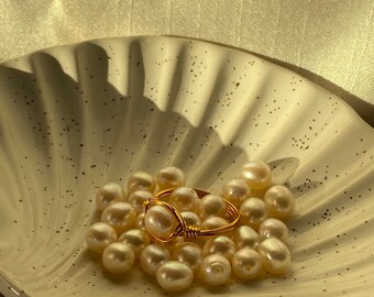 May include: A gold ring with a pearl wrapped around it, surrounded by a collection of white pearls in a white ceramic shell.