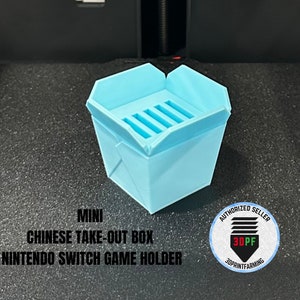 May include: A light blue 3D printed mini Chinese takeout box game holder for Nintendo Switch games. The box has a slot for the game cartridge and says "MINI CHINESE TAKE-OUT BOX NINTENDO SWITCH GAME HOLDER" on the front. The image also includes a round sticker with the text "AUTHORIZED SELLER 3DPF 3DPRINTFARMING".
