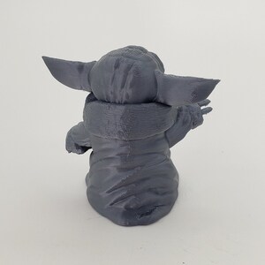 May include: A gray 3D printed figurine of a character from the Star Wars franchise, known as "The Child" or "Baby Yoda". The figurine is sitting with its arms raised and its head tilted back.