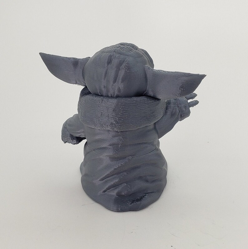 May include: A gray 3D printed figurine of a character from the Star Wars franchise, known as "The Child" or "Baby Yoda". The figurine is sitting with its arms raised and its head tilted back.