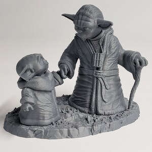 Peut inclure: Une figurine en 3D imprim&eacute;e en gris de deux personnages Yoda de Star Wars. Le plus grand Yoda tient un b&acirc;ton de marche et le plus petit Yoda tient la main du plus grand Yoda. Les figurines sont debout sur une base grise.