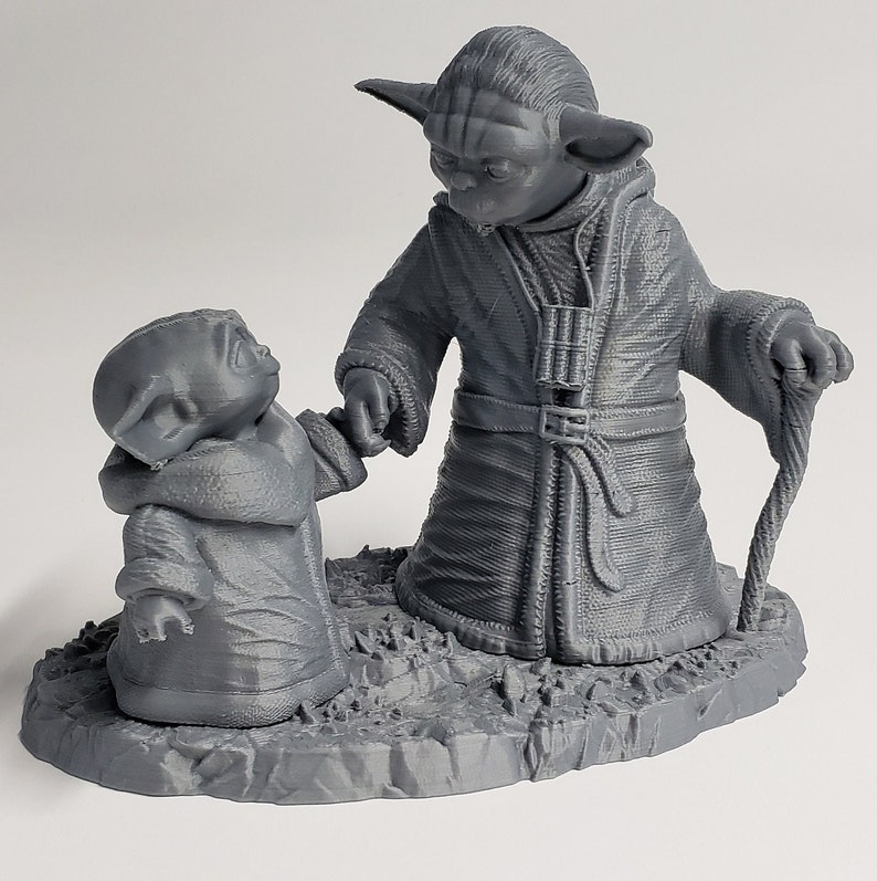 Peut inclure: Une figurine en 3D imprim&eacute;e en gris de deux personnages Yoda de Star Wars. Le plus grand Yoda tient un b&acirc;ton de marche et le plus petit Yoda tient la main du plus grand Yoda. Les figurines sont debout sur une base grise.
