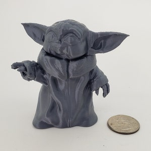 May include: A gray 3D printed figurine of a character from the Star Wars franchise, known as "The Child" or "Baby Yoda". The figurine is standing on a white surface and is about the size of a quarter.