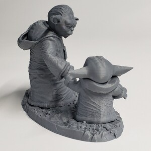 Peut inclure: Une figurine en 3D imprim&eacute;e en gris de Yoda et Grogu de Star Wars. Yoda est debout, les bras tendus, et Grogu est debout devant lui, regardant Yoda. La figurine est pos&eacute;e sur une base grise.