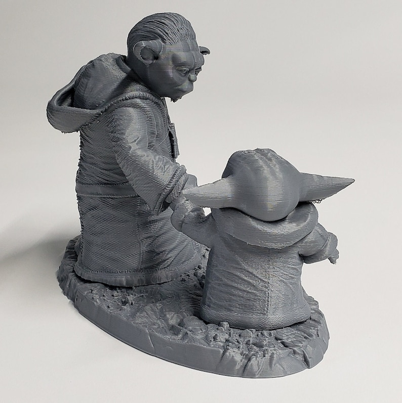 Peut inclure: Une figurine en 3D imprim&eacute;e en gris de Yoda et Grogu de Star Wars. Yoda est debout, les bras tendus, et Grogu est debout devant lui, regardant Yoda. La figurine est pos&eacute;e sur une base grise.