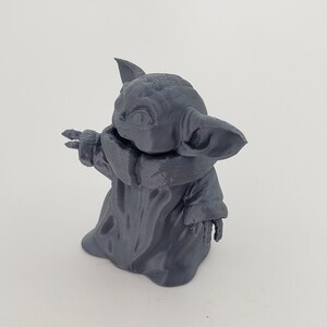 May include: A gray 3D printed figurine of a character from the Star Wars franchise, known as "The Child" or "Baby Yoda". The figurine is standing with its arms outstretched, and its head is tilted to the side.