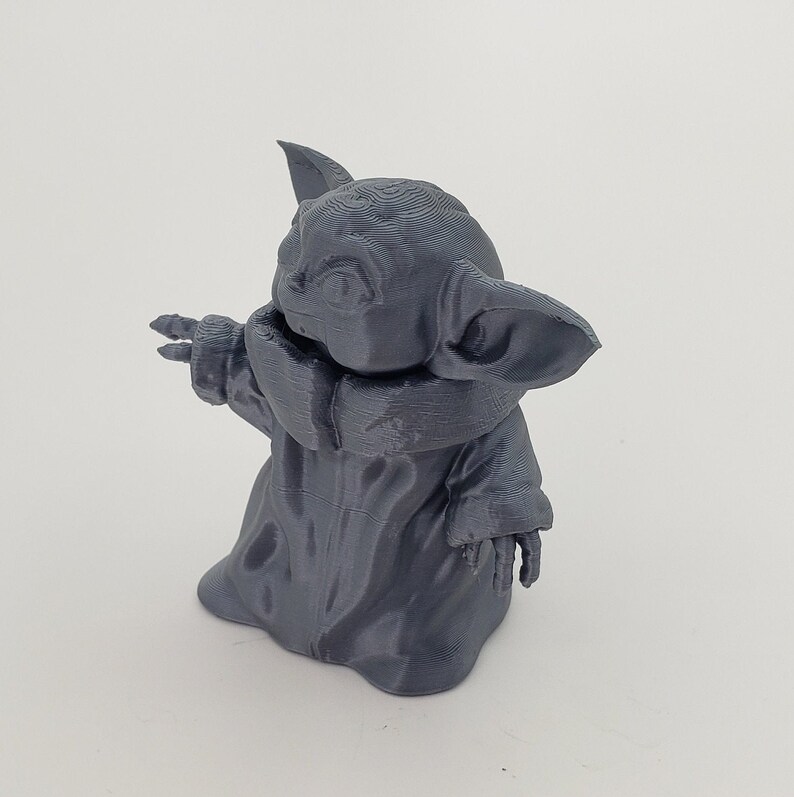 May include: A gray 3D printed figurine of a character from the Star Wars franchise, known as "The Child" or "Baby Yoda". The figurine is standing with its arms outstretched, and its head is tilted to the side.