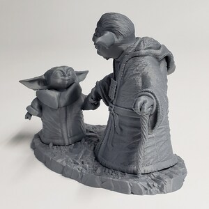 Peut inclure: Une figurine en 3D imprim&eacute;e en gris de Yoda et un jeune Yoda, debout sur une base grise. Les figurines se tiennent la main et portent toutes les deux des robes.