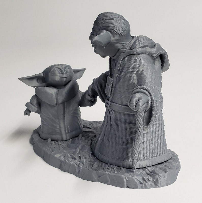 Peut inclure: Une figurine en 3D imprim&eacute;e en gris de Yoda et un jeune Yoda, debout sur une base grise. Les figurines se tiennent la main et portent toutes les deux des robes.
