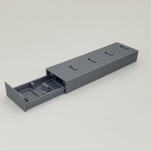 May include: A grey rectangular storage box with a sliding drawer. The box has four rectangular cutouts on the top. The drawer is partially extended, revealing compartments inside. The item is made of a solid, matte material.