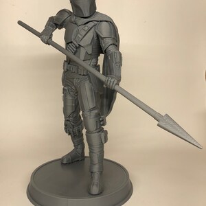 Puede incluir: Una figura de acci&oacute;n gris impresa en 3D de un guerrero mandaloriano sosteniendo una lanza. La figura est&aacute; de pie sobre una base redonda.