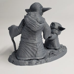Peut inclure: Une figurine en 3D imprim&eacute;e en gris de Yoda et un plus petit Yoda tenant un b&acirc;ton. Le plus grand Yoda porte une robe et tient un b&acirc;ton dans sa main. Les figurines sont debout sur une base grise.