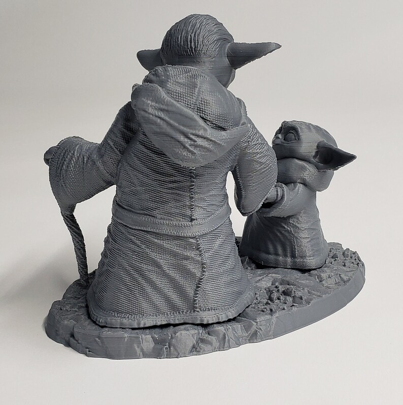 Peut inclure: Une figurine en 3D imprim&eacute;e en gris de Yoda et un plus petit Yoda tenant un b&acirc;ton. Le plus grand Yoda porte une robe et tient un b&acirc;ton dans sa main. Les figurines sont debout sur une base grise.