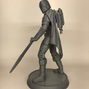 Puede incluir: Una figura de acci&oacute;n gris impresa en 3D de un personaje con casco y sosteniendo un arma. La figura est&aacute; de pie sobre una base redonda.