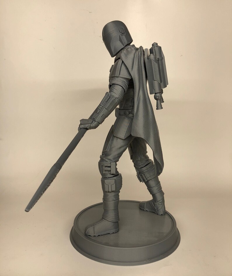 Puede incluir: Una figura de acci&oacute;n gris impresa en 3D de un personaje con casco y sosteniendo un arma. La figura est&aacute; de pie sobre una base redonda.