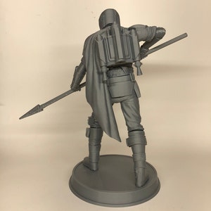 Puede incluir: Una figura gris impresa en 3D de un personaje del universo de Star Wars, sosteniendo una lanza. El personaje lleva un casco y una armadura, y tiene una mochila propulsora en la espalda.