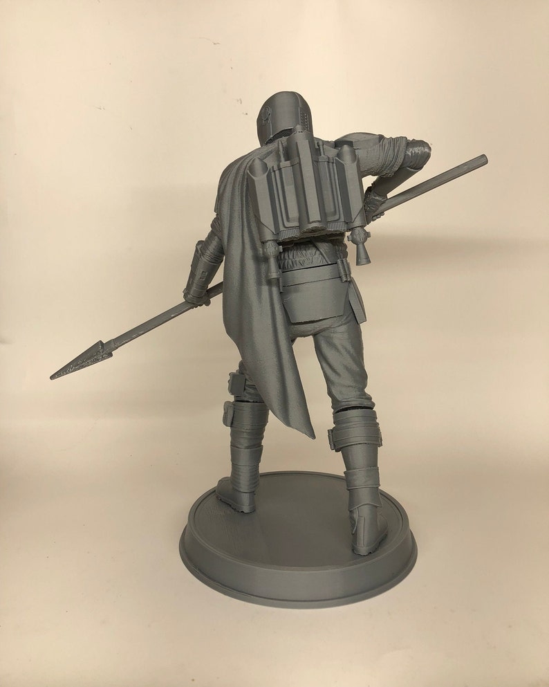 Puede incluir: Una figura gris impresa en 3D de un personaje del universo de Star Wars, sosteniendo una lanza. El personaje lleva un casco y una armadura, y tiene una mochila propulsora en la espalda.
