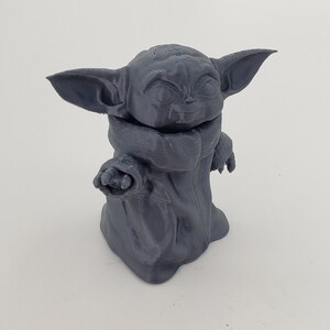 May include: A gray 3D printed figurine of a character from the Star Wars franchise, known as "The Child" or "Baby Yoda". The figurine is standing with its arms outstretched and has a large head with big eyes and a small mouth.