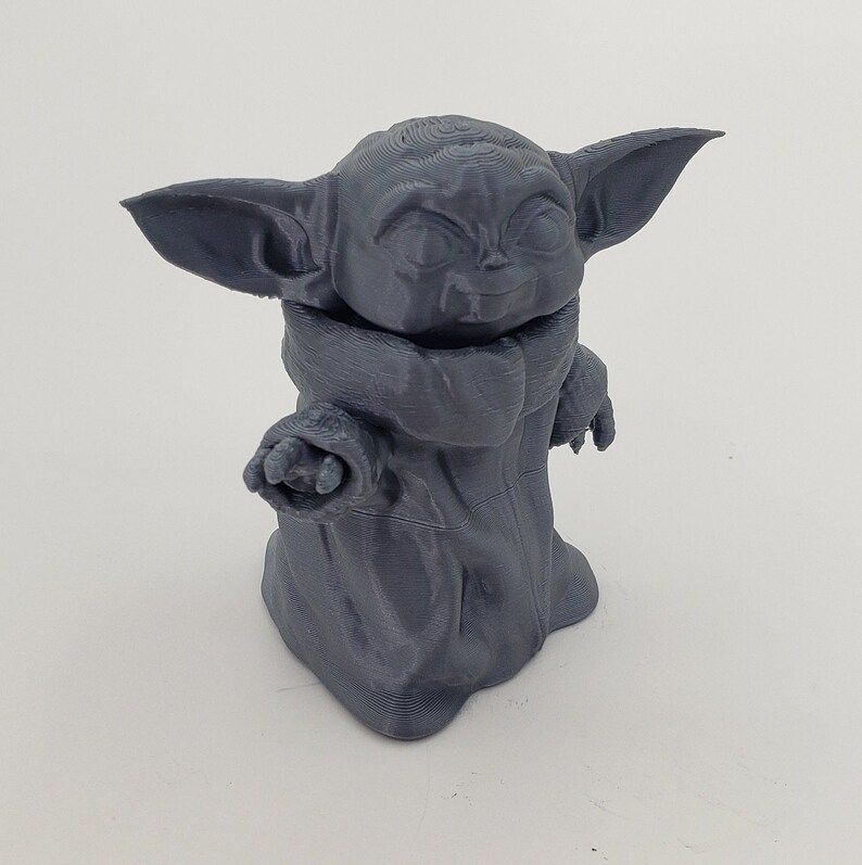 May include: A gray 3D printed figurine of a character from the Star Wars franchise, known as "The Child" or "Baby Yoda". The figurine is standing with its arms outstretched and has a large head with big eyes and a small mouth.