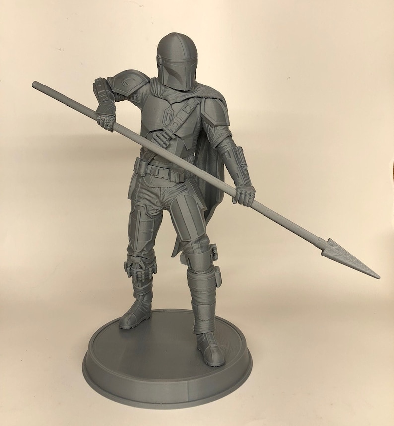 Puede incluir: Una figura gris de un guerrero mandaloriano sosteniendo una lanza, impresa en 3D. La figura est&aacute; de pie sobre una base redonda.
