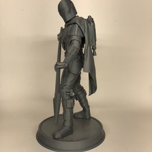 Puede incluir: Una figura de acci&oacute;n gris impresa en 3D de un personaje del universo de Star Wars. La figura sostiene un arma y lleva un casco y una capa. La figura est&aacute; de pie sobre una base redonda.