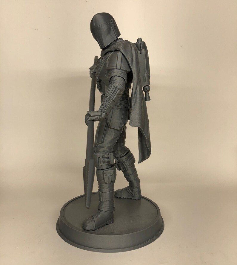 Puede incluir: Una figura de acci&oacute;n gris impresa en 3D de un personaje del universo de Star Wars. La figura sostiene un arma y lleva un casco y una capa. La figura est&aacute; de pie sobre una base redonda.