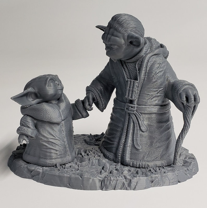 Peut inclure: Une figurine en 3D imprim&eacute;e en gris de Yoda et un plus petit Yoda, tous deux avec une peau verte et de grandes oreilles. Le plus grand Yoda tient un b&acirc;ton et porte une robe. Le plus petit Yoda tient la main du plus grand Yoda. Les figurines sont debout sur une base grise.