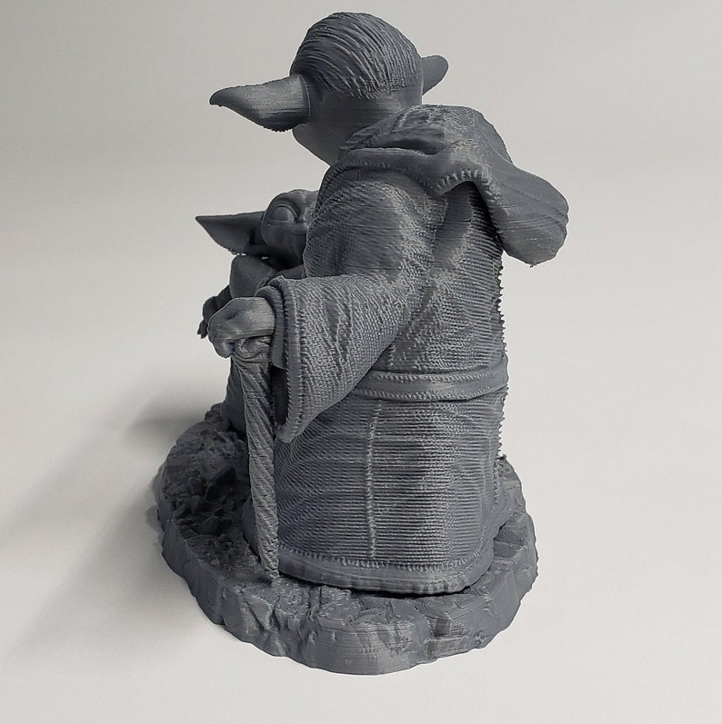 Peut inclure: Une figurine en plastique gris imprim&eacute;e en 3D du personnage de Star Wars, Yoda, tenant un b&acirc;ton de marche. Yoda porte une robe et a une petite figurine verte de Baby Yoda assise sur ses genoux.