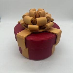 Puede incluir: Una caja de regalo redonda roja con un lazo marrón en la parte superior. El lazo está hecho de múltiples bucles y se asienta sobre la caja. Cintas marrones envuelven los lados de la caja. La caja es de color sólido y se encuentra sobre una superficie blanca.