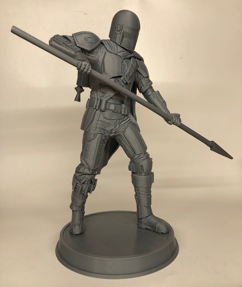 Puede incluir: Una figura gris impresa en 3D de un personaje del universo de Star Wars, sosteniendo una lanza. La figura est&aacute; de pie sobre una base redonda.