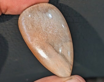 May include: A teardrop-shaped sunstone cabochon with a light brown and white color. The stone has a shimmery, iridescent quality.