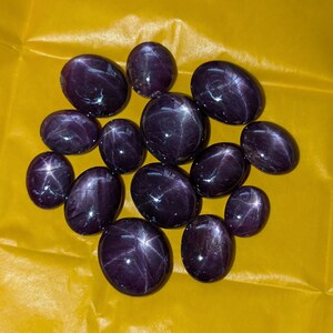 Puede incluir: Una colección de piedras preciosas de rubí estrella, de forma ovalada y color púrpura oscuro. Cada piedra muestra un efecto de estrella prominente, con líneas que irradian desde un punto central. Las gemas están dispuestas sobre una superficie amarilla brillante.