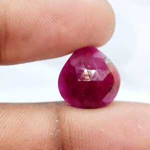 Puede incluir: Una gema facetada de color rojo intenso, sostenida entre dos dedos. La gema tiene forma de pera con múltiples facetas que reflejan la luz. El color es un rojo intenso y saturado, con una ligera variación de tono.