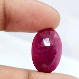 Puede incluir: Una gema de rubí facetada de forma ovalada. El rubí es de un color rojo intenso y rico, con una superficie pulida y reflectante. La gema se sostiene entre dos dedos sobre un fondo blanco.