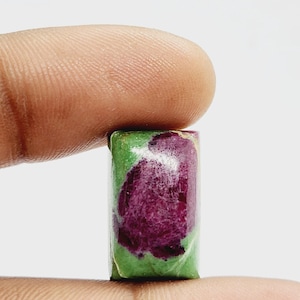 May include: A rectangular gemstone with a vibrant mix of colours. The stone features a deep magenta centre surrounded by a green border. The gemstone is held between two fingers, showcasing its polished surface.