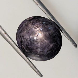 Puede incluir: Una piedra preciosa redonda de color púrpura oscuro con un patrón en forma de estrella, sostenida por unas pinzas.