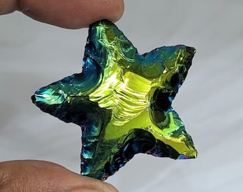 May include: A star-shaped piece of iridescent obsidian with a glossy, reflective surface. The stone displays a vibrant spectrum of colors, shifting from deep blue and purple tones at the edges to a bright, luminous lime green in the center.