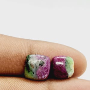 Peut inclure: Deux pierres précieuses carrées avec un mélange de couleurs. Les pierres précieuses présentent un mélange vibrant de teintes roses, vertes et blanches. Les pierres précieuses sont tenues dans une main, avec un fond blanc.