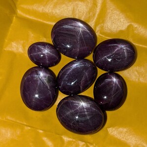 Puede incluir: Siete piedras preciosas ovaladas y pulidas con un tono púrpura intenso están dispuestas sobre una superficie amarilla. Cada piedra muestra un patrón en forma de estrella en su superficie. Las piedras preciosas varían ligeramente en tamaño y forma, con un acabado brillante.