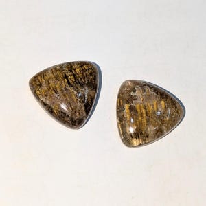 Op de afbeelding: Twee driehoekige edelstenen met bruine, gouden en heldere tinten. De gepolijste stenen tonen natuurlijke patronen en texturen. De edelstenen zijn geplaatst tegen een witte achtergrond. Ideaal voor sieraden.