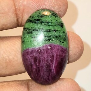 May include: An oval-shaped cabochon gemstone featuring a distinct colour split. The upper portion is a vibrant green with dark black inclusions, while the lower portion is a deep, saturated magenta-purple with a crystalline, textured appearance.
